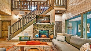 A cozy log cabin interior with a stone fireplace, a wooden staircase, and a large window. - Generational Cabin Near Gatlinburg & Dollywood (Sevierville)