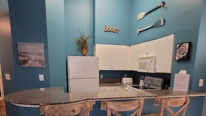 A modern kitchen with blue walls, wooden chairs, a white refrigerator, and a microwave. - Gorgeous Ground Floor Condo! (Panama City Beach)