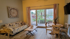 A living room with a sofa, coffee table, and chairs, a TV mounted on the wall, and a balcony with a view of buildings and greenery. - Gorgeous Ground Floor Condo! (Panama City Beach)