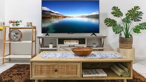 Ein Wohnzimmer mit einem Flachbildfernseher auf einem Holzständer, einem Korbwäscher auf einem Couchtisch, einer Kübelpflanze mit einem Monstera und einem Regal mit Ventilator und Dekoration.