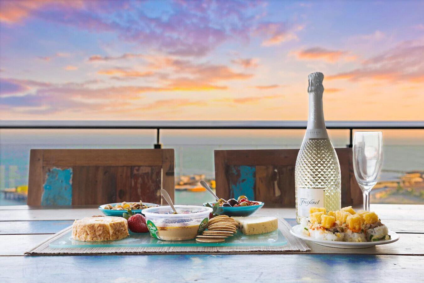 Une table dressée avec divers plats et une bouteille de vin mousseux Freixenet, le tout mis en valeur par une vue panoramique sur l’océan.