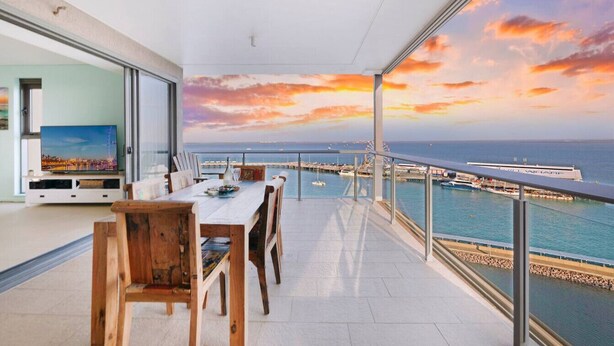 Ein Balkon mit Blick auf das Meer und ein Riesenrad, ein Esstisch mit Stühlen und ein Fernseher im Hintergrund.
