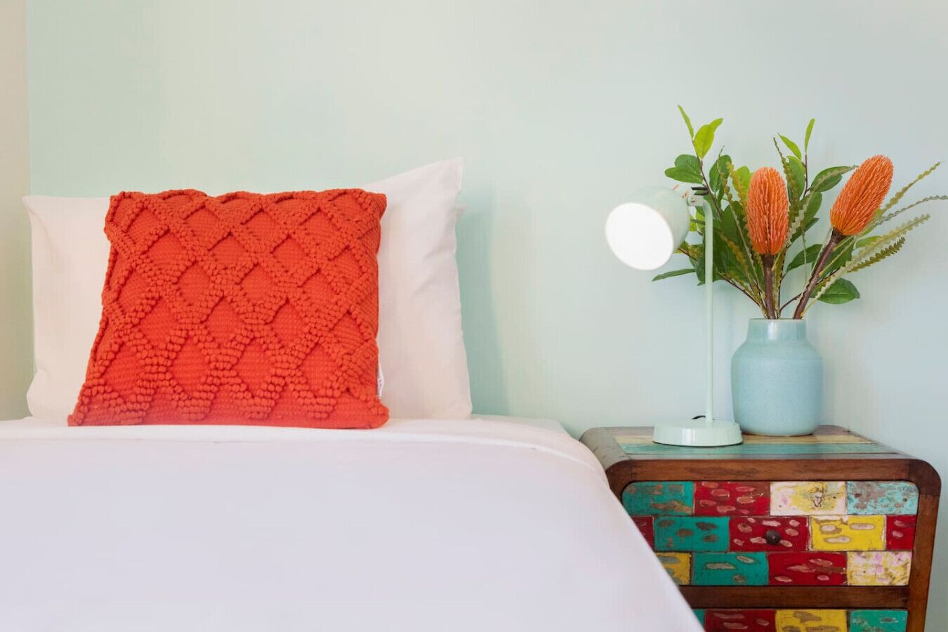 Una cama con un almohadón rojo texturizado, un almohadón blanco y una mesita de noche colorida de retazos con un jarrón de flores naranjas.
