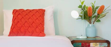 Una cama con un almohadón rojo texturizado, un almohadón blanco y una mesita de noche colorida de retazos con un jarrón de flores naranjas.