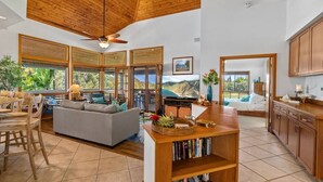 A spacious living area with a wooden ceiling, large windows, a grey sofa, and a wooden bookshelf.