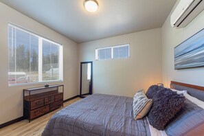 A bedroom with a large bed, a dresser, two windows with blinds, a ceiling light, and an air conditioning unit.