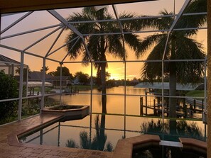 Blick auf einen Pool mit Glasumrandung, Palmen und einem Sonnenuntergang im Hintergrund.