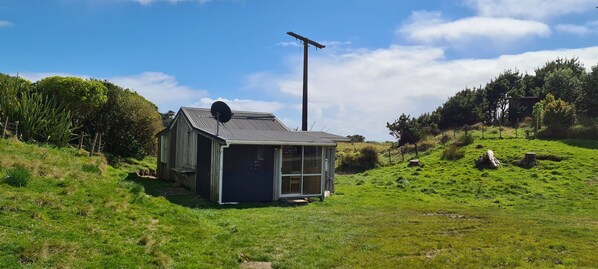 Une petite maison à un seul étage, avec un toit en métal et une antenne parabolique, entourée d’un paysage verdoyant et d’arbres.