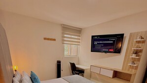 Et soveværelse med seng, natbord, lampe, skrivebord, stol, fjernsyn og en reol med pynteting.