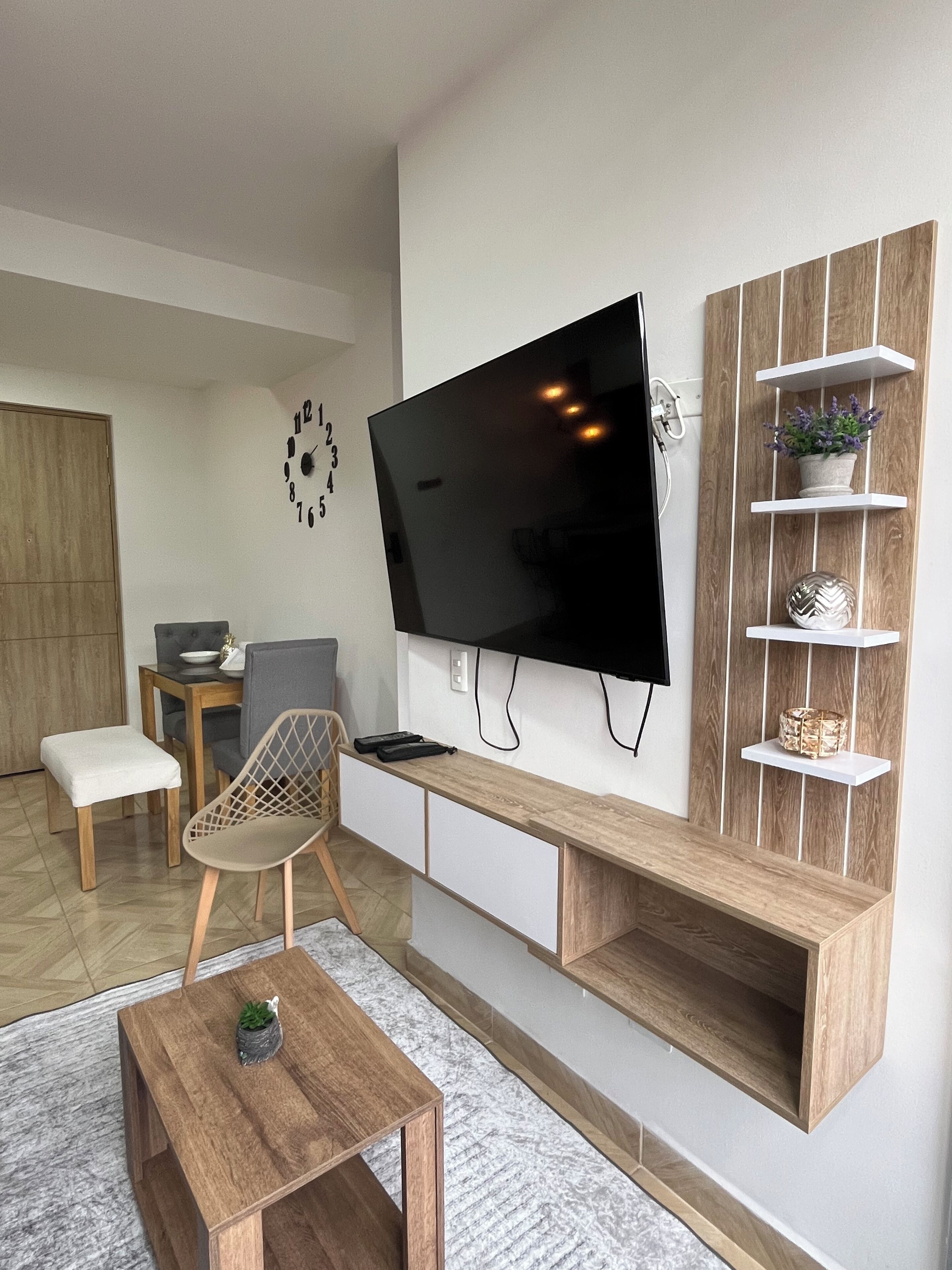 A modern living room with a flat-screen TV mounted on the wall, wooden shelves, a grey chair, a small table with a plant, and a wooden bench.