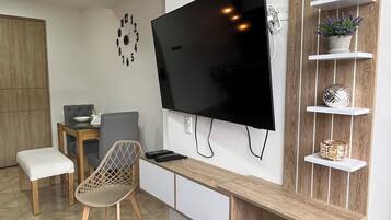 A modern living room with a flat-screen TV mounted on the wall, wooden shelves, a grey chair, a small table with a plant, and a wooden bench.