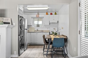 A compact kitchen with white cabinetry, a stainless steel refrigerator, a dining table with chairs, and a window with blinds.