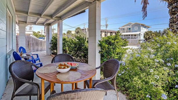 A covered patio with wicker furniture and a table set with food, surrounded by greenery and buildings in the background. - South Beach Cabana Down | Patio | Pool | Across Street from Beach (Fernandina Beach)