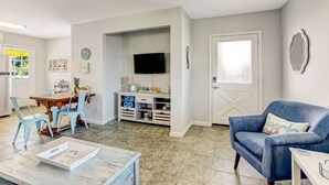 A modern living room with a blue sofa, a wooden dining table with chairs, a TV, and a bookshelf. - South Beach Cabana Down | Patio | Pool | Across Street from Beach (Fernandina Beach)