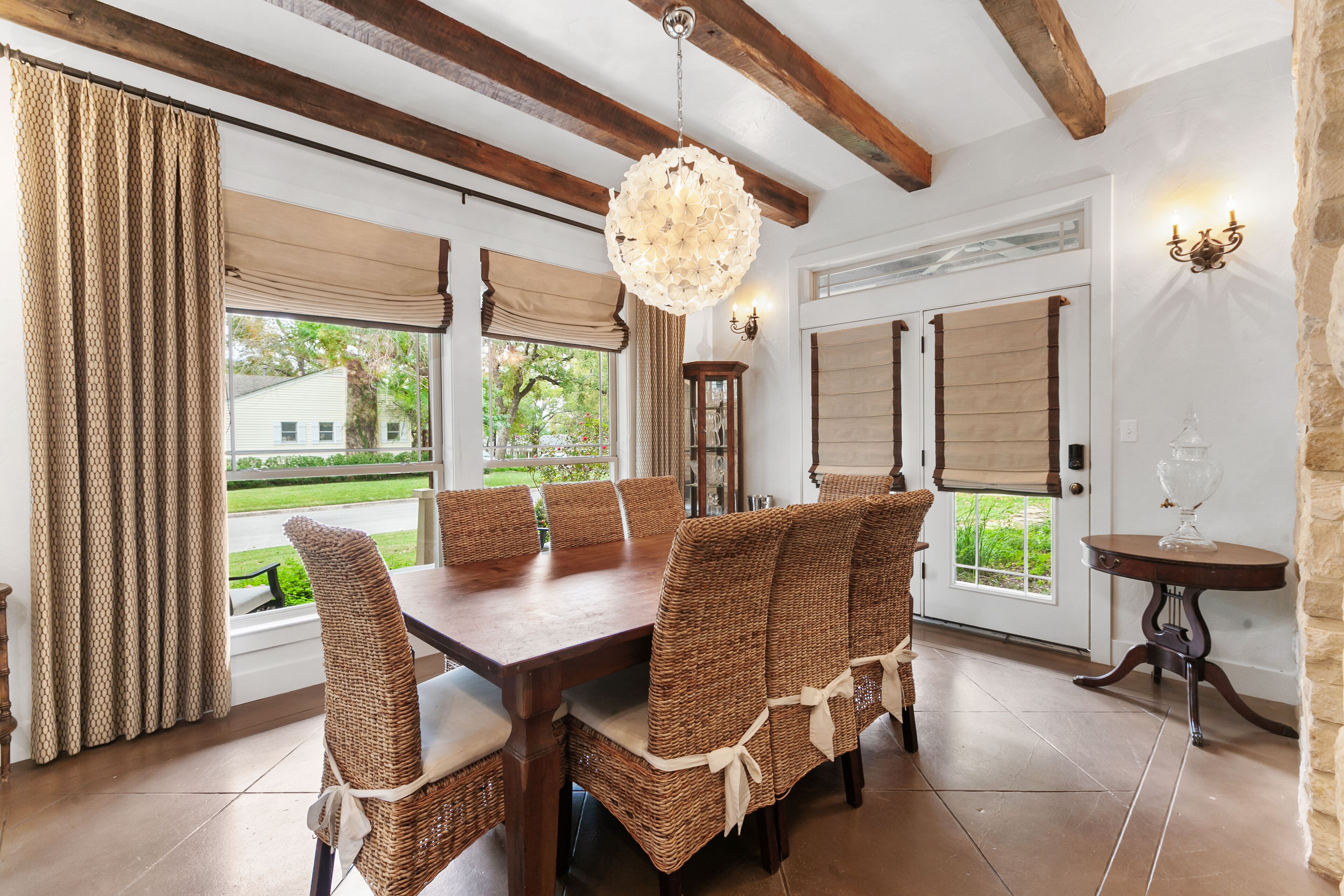 Un comedor con mesa de madera y sillas de mimbre, ventanas amplias con cortinas y un candelabro.