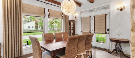 Un comedor con mesa de madera y sillas de mimbre, ventanas amplias con cortinas y un candelabro.