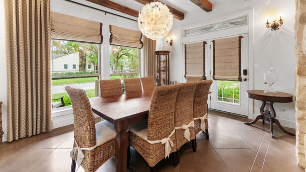 A dining area with a wooden table and wicker chairs, large windows with curtains, and a chandelier.