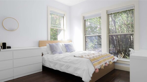 A bedroom with a bed, white walls, a wooden headboard, and large windows with a view of trees.