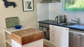 A modern kitchen with white cabinetry, a stainless steel range hood, a built-in oven, and a dishwasher. There is a window providing a view of greenery outside.