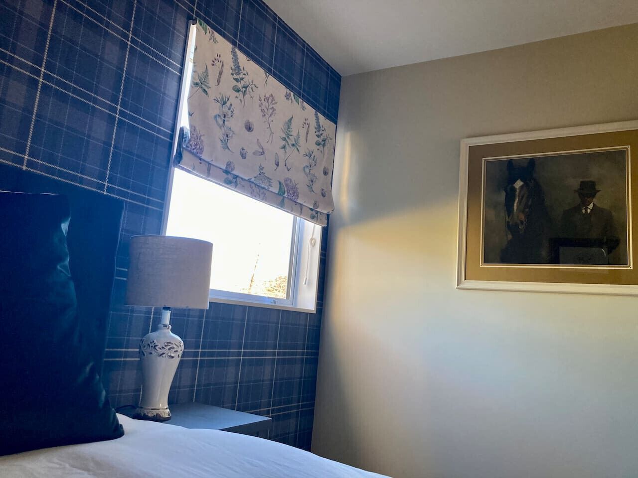 Una camera d'albergo con un letto, un comodino con una lampada, una finestra con tenda a fiori e un quadro appeso al muro.