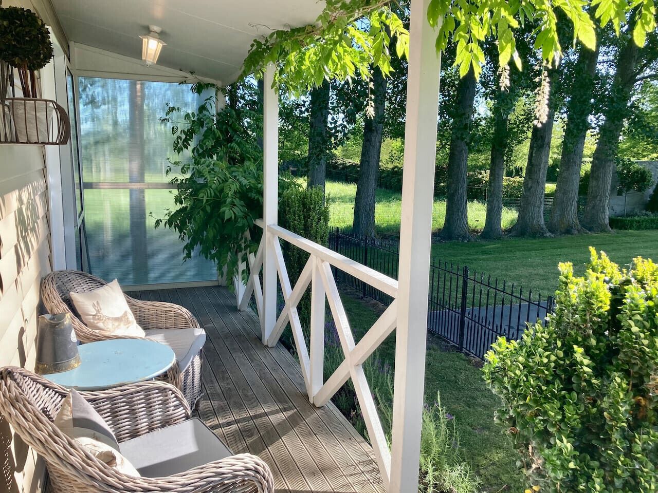 A covered outdoor seating area with wicker furniture and a table, surrounded by greenery and a fence.
