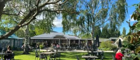 An outdoor seating area with tables and chairs, a building with a roof, and trees in the background.