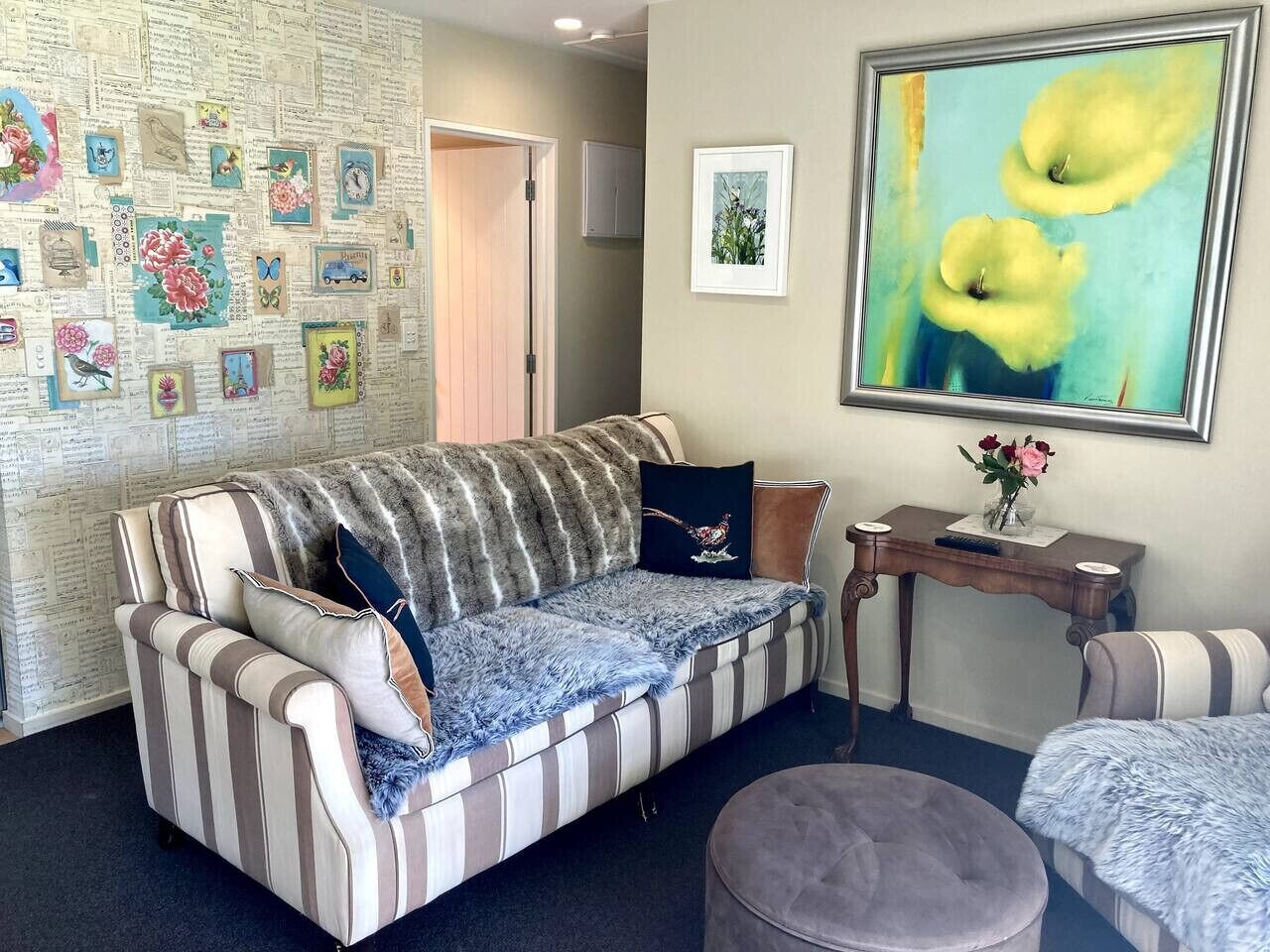 A living room with a striped sofa, a round ottoman, a small table with a vase, and a wall decorated with various framed pictures.