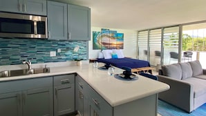 A modern kitchen with a built-in microwave, a sink, and a decorative backsplash. There is a bed with a blue bedspread and pillows, and a sofa with cushions. A large window provides a view of the outdoors. - Beachfront & Oceanviews - New Furnishings (Hauula)