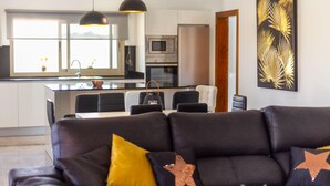 Una sala de estar moderna con un sofá oscuro, una mesa de centro y un comedor con sillas. Se ve una cocina al fondo con armarios blancos y un microondas.