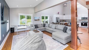 A spacious living room with a large sectional sofa, a coffee table, and a foosball table. There is a kitchen area with modern appliances and a dining table.