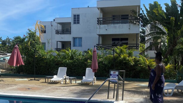 A swimming pool with lounge chairs and umbrellas, surrounded by greenery and a multi-story building in the background.