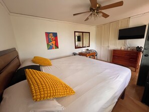 Ein Schlafzimmer mit einem Bett, einem Deckenventilator, einem Schreibtisch mit Stuhl, einem Kleiderschrank und einem an der Wand befestigten Fernseher.