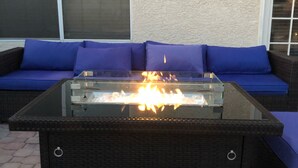 Una mesa con cubierta de vidrio y chimenea al aire libre, rodeada por un sofá de mimbre oscuro con cojines azules, ubicada en un patio con baldosas hexagonales.