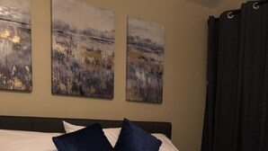 Una cama bien hecha con ropa de cama blanca y dos almohadas azules, una cesta tejida con toallas dobladas, y tres cuadros abstractos en la pared.