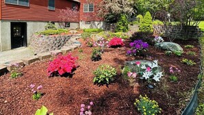 Una casa de madera roja con un muro de piedra de contención y un jardín con diversas plantas florecientes.