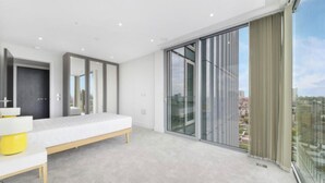 Ein modernes Schlafzimmer mit einem großen Bett, einer Bank und Blick auf die Stadt durch raumhohe Fenster.