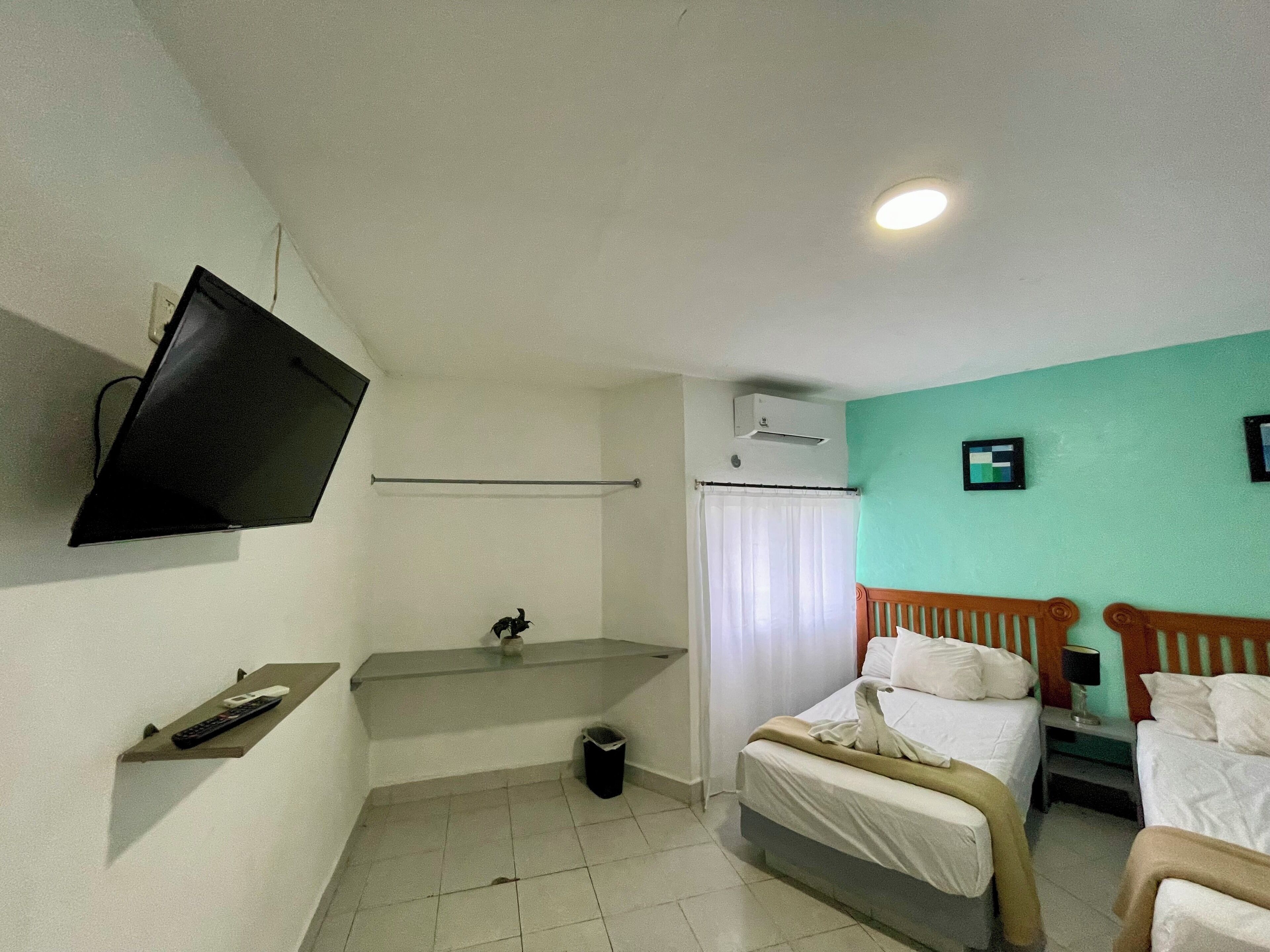 A hotel room with a bed, a TV mounted on the wall, a small shelf with a remote control, a trash bin, and an air conditioning unit.