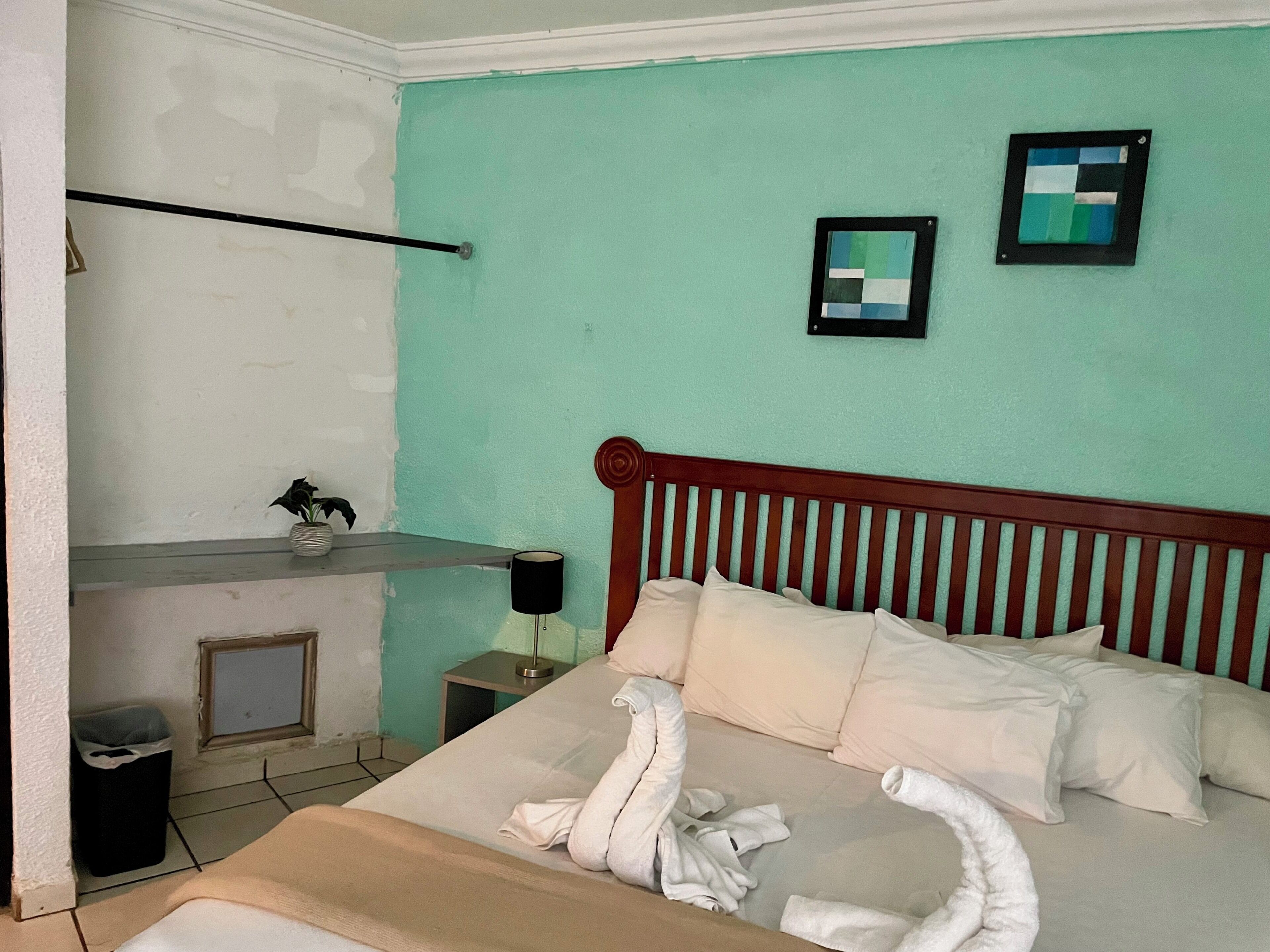 A bedroom with a bed, a bedside table, a lamp, a framed picture, and a wall with a partial view of a window.