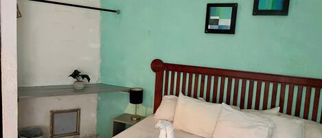 A bedroom with a bed, a bedside table, a lamp, a framed picture, and a wall with a partial view of a window.