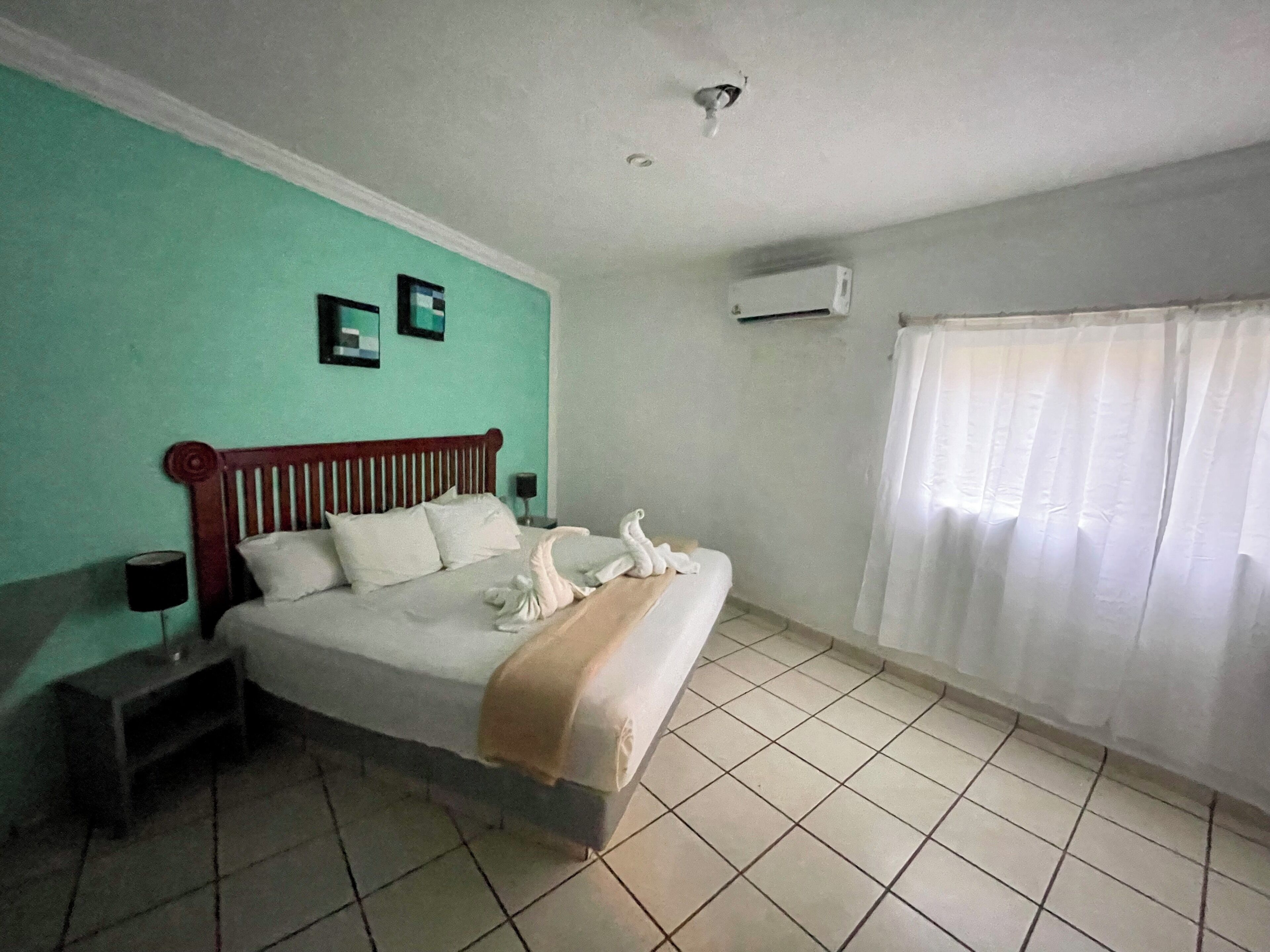 A hotel room with a bed, a nightstand, a wall-mounted air conditioner, and a window with curtains.
