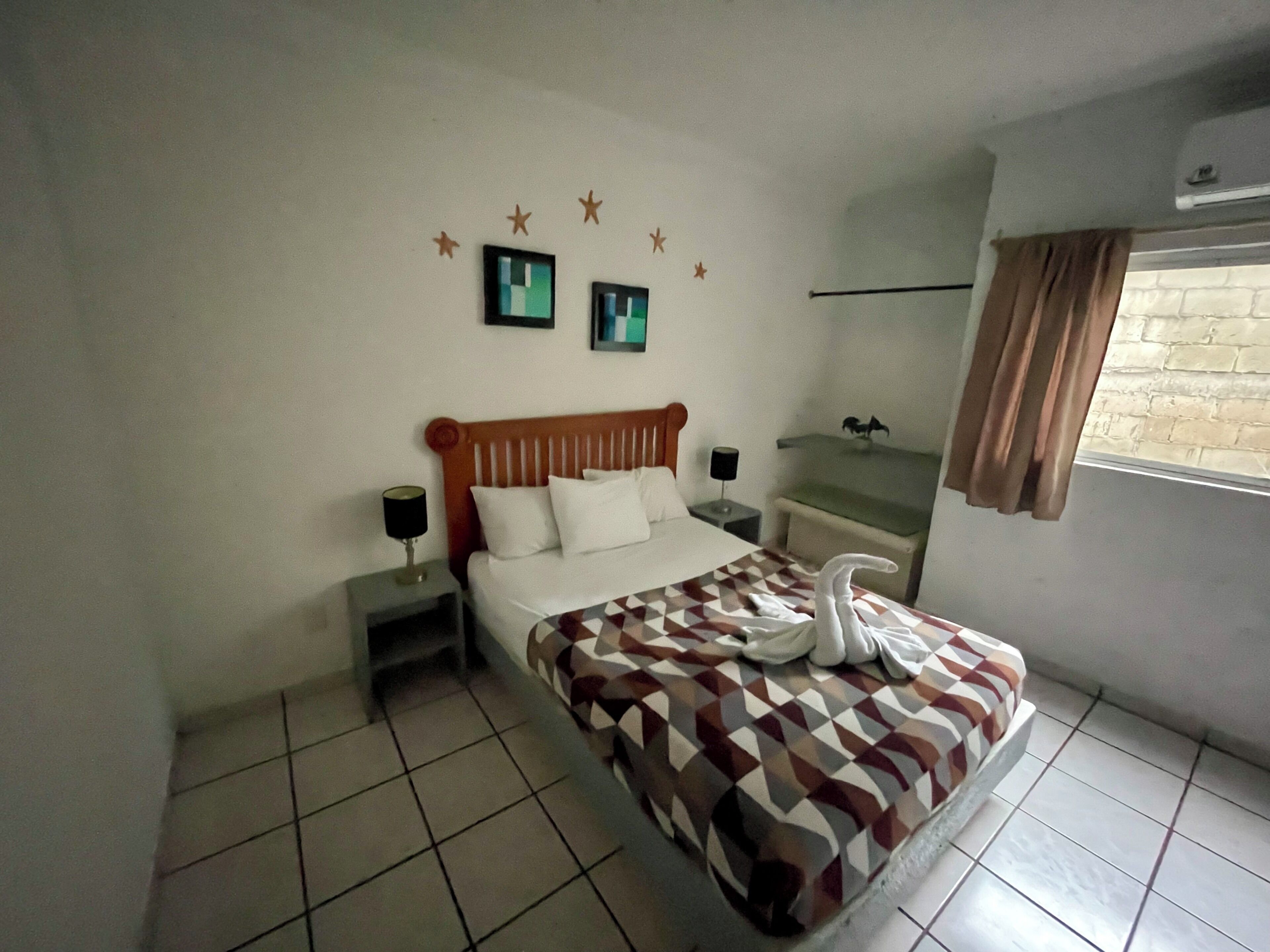 Quarto de hotel com cama, mesa de cabeceira, ar condicionado fixado na parede e janela com cortinas.