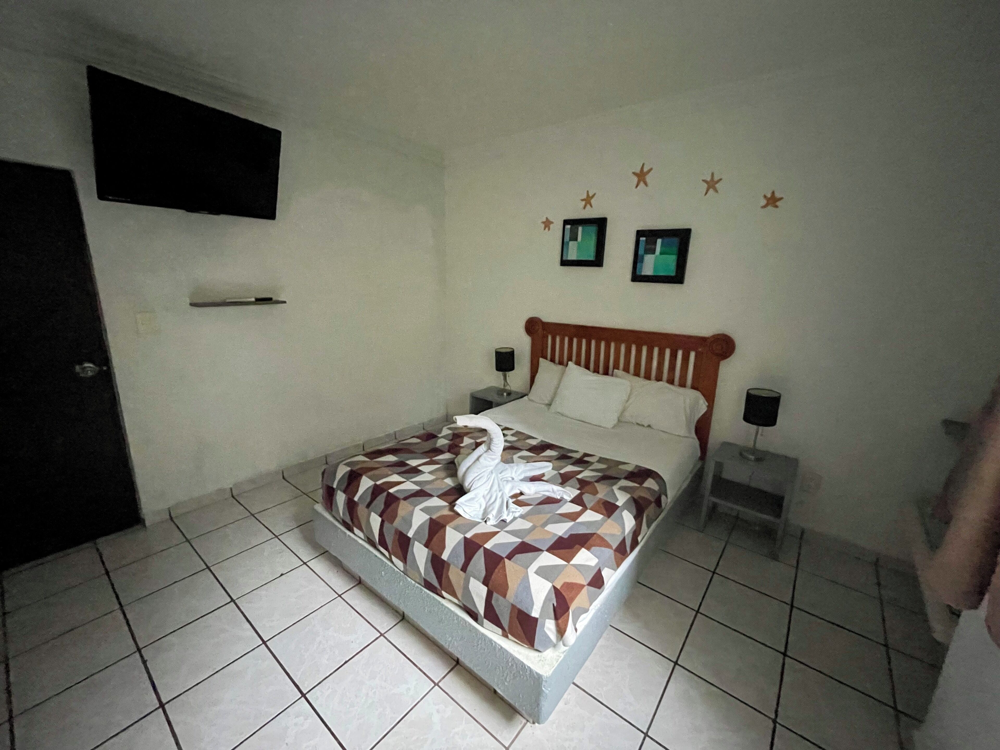 Una cama bien hecha con una colcha de estampado geométrico, dos mesitas de noche con lámparas, un televisor de pantalla plana montado en la pared y una puerta a la izquierda.