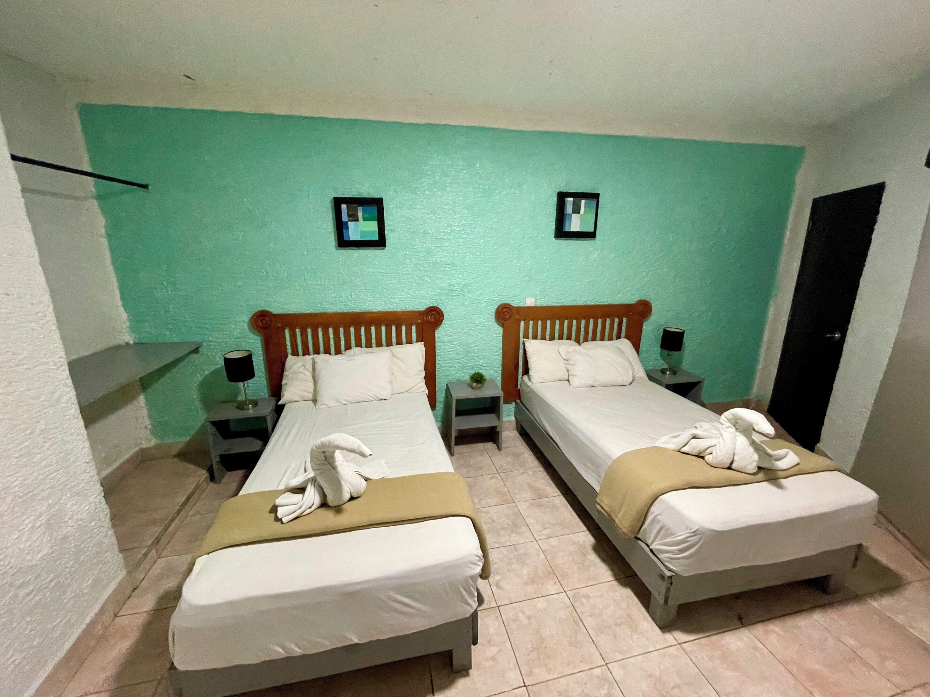 A twin room with two beds, each with a wooden headboard, white bedding, and a towel on the beds. There are two bedside tables with lamps and a small plant on one. The walls are painted teal, and there are two framed pictures on the wall.