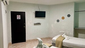 Una habitación de hotel con dos camas, un televisor montado en la pared, una puerta con un cartel de 'no fumar' y una pequeña estantería.