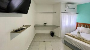Una habitación pequeña y limpia con cama, televisor montado en la pared, estante pequeño, toallero y basurero.