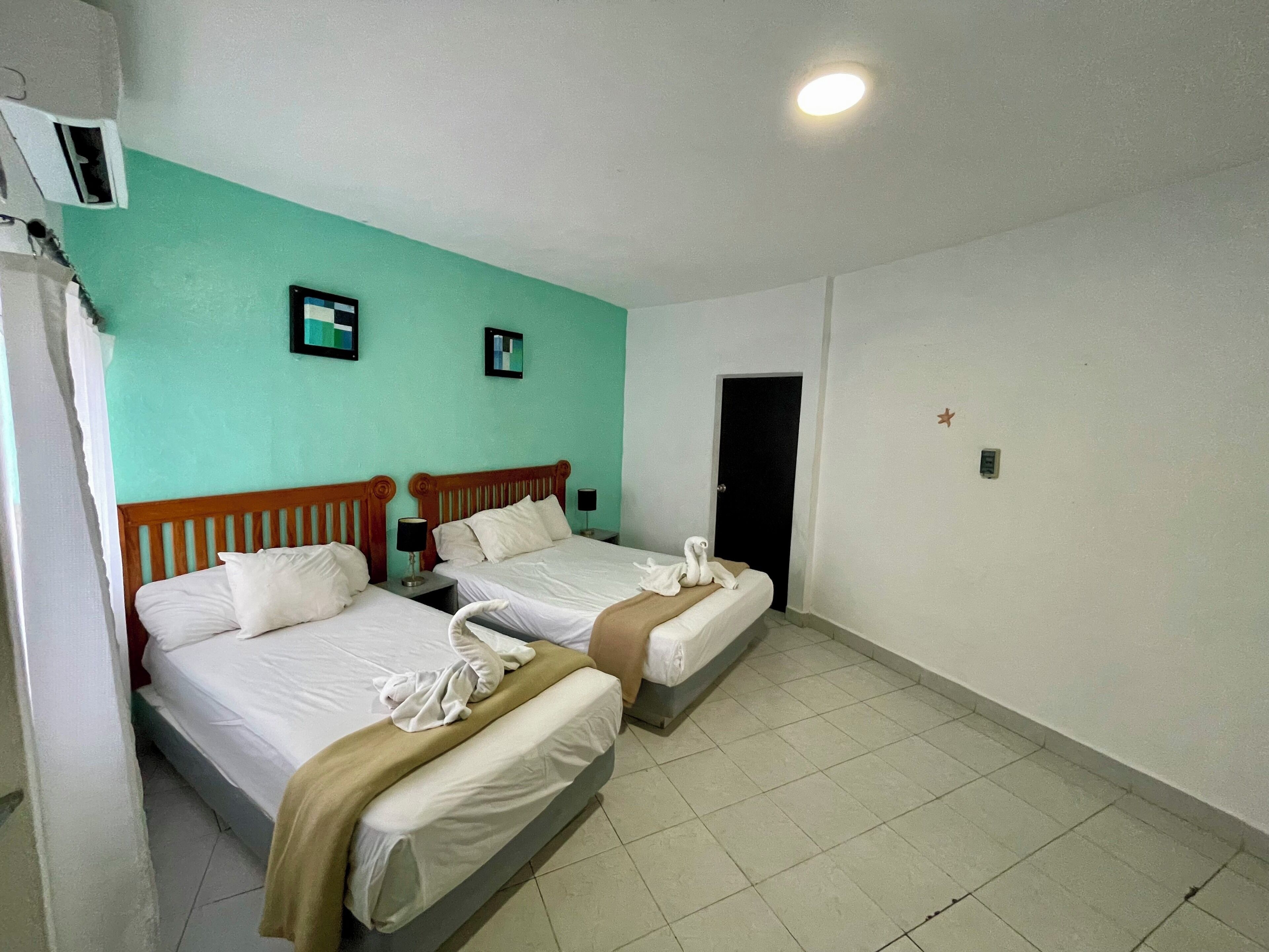 A hotel room with two beds, a teal-colored wall, a door, and a ceiling light.