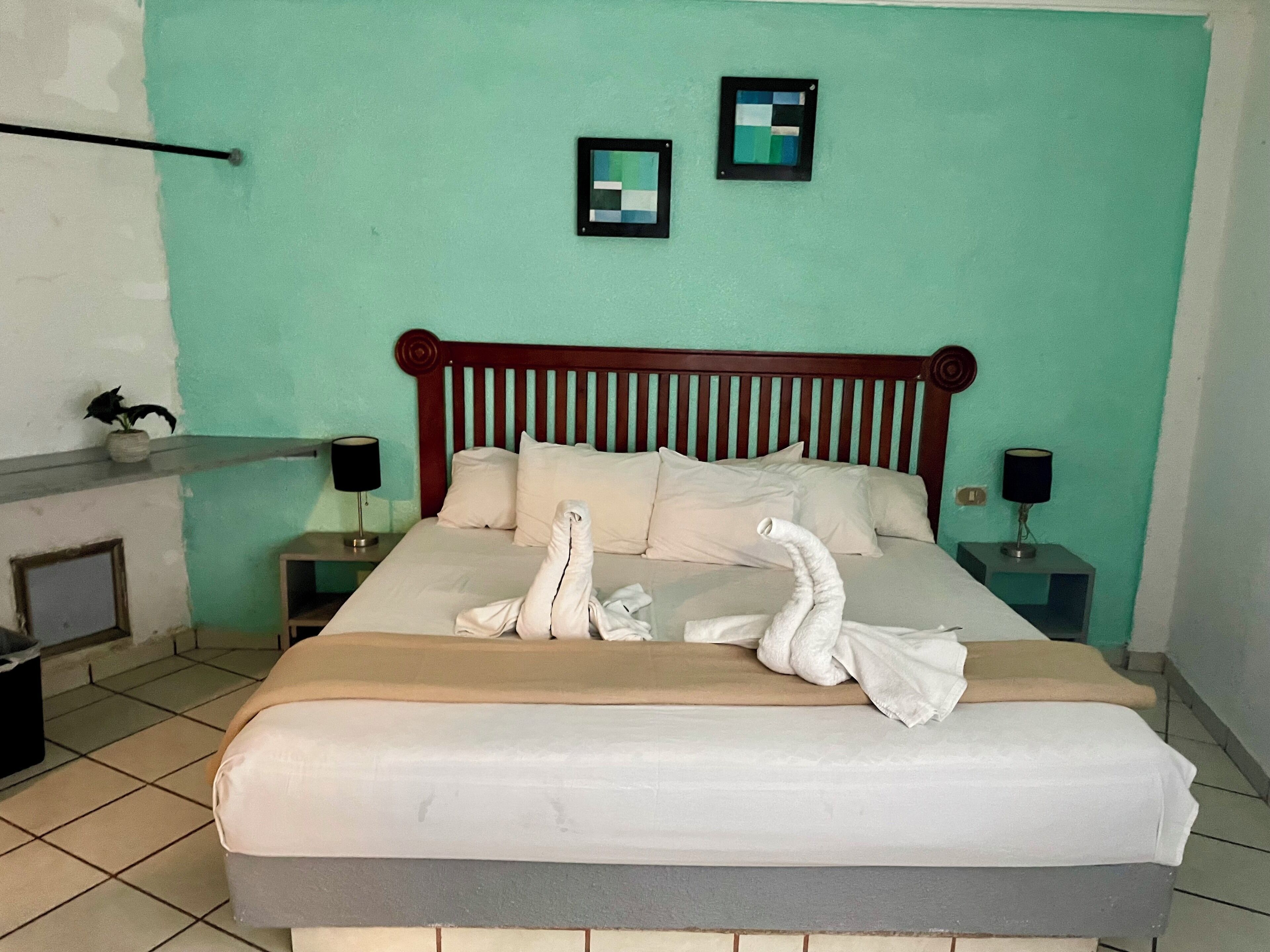 A neatly made bed with white linens and two decorative swans, a wooden headboard, two wall-mounted picture frames, and bedside lamps on a teal-colored wall.