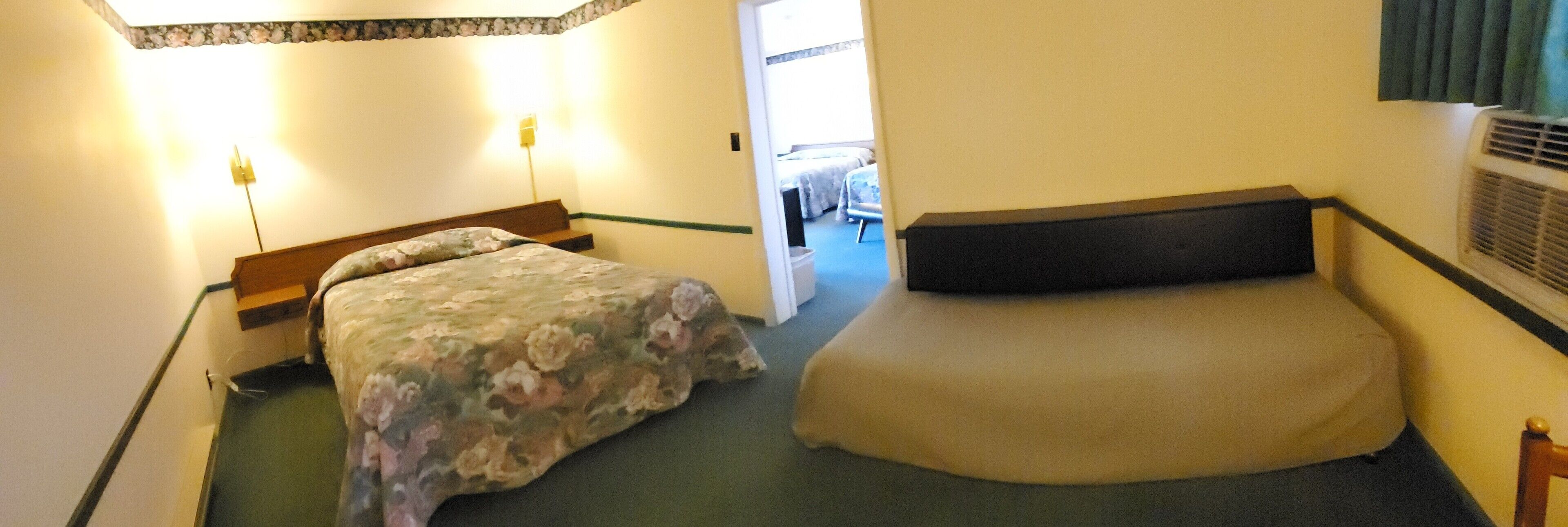 A small hotel room with two beds, a wooden headboard, a bedside table, a lamp, and a window with curtains.