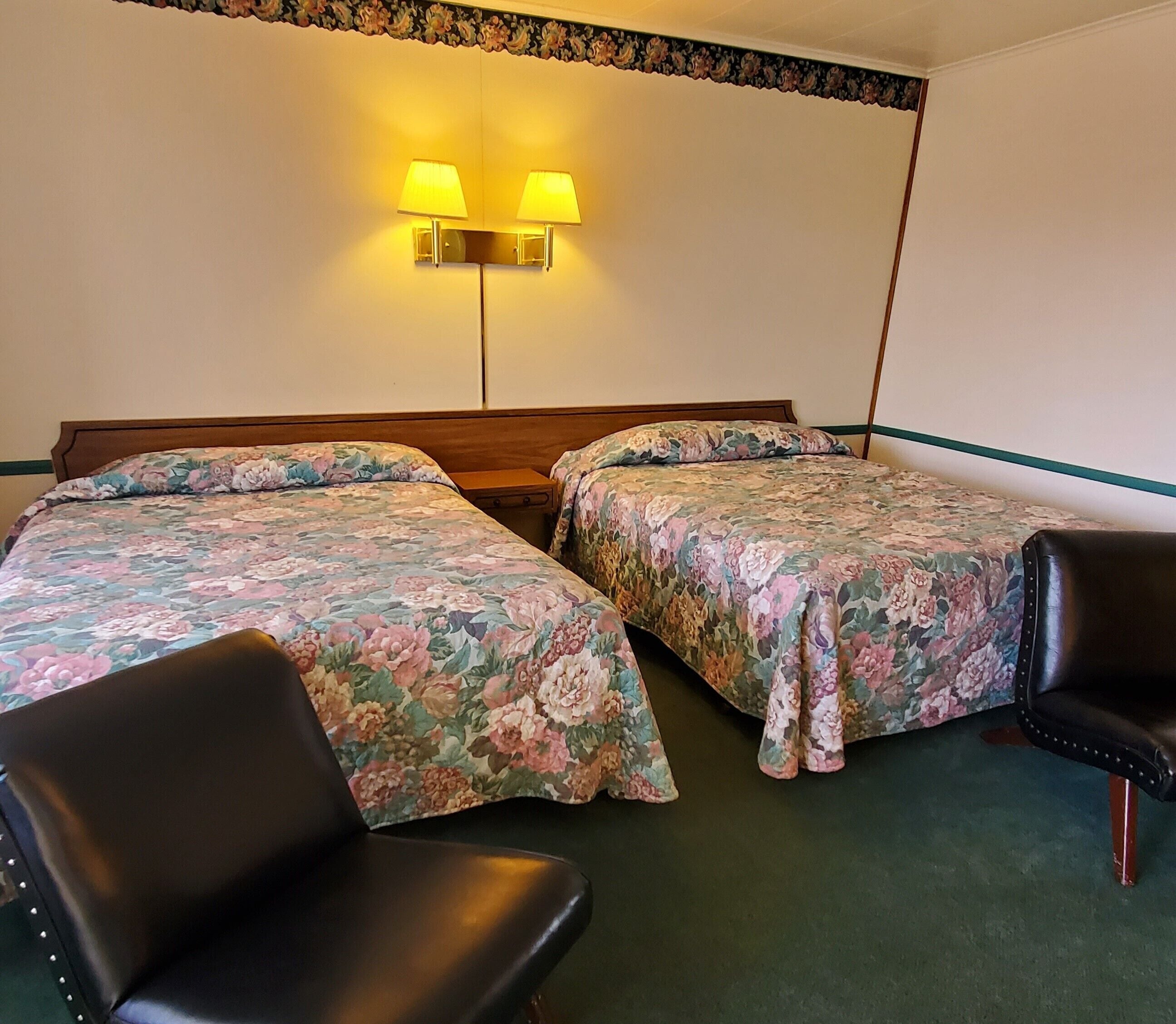 Une chambre d’hôtel avec deux lits, chacun agrémenté d’une literie à motifs floraux, une table de chevet, une chaise et des lampes fixées au mur.