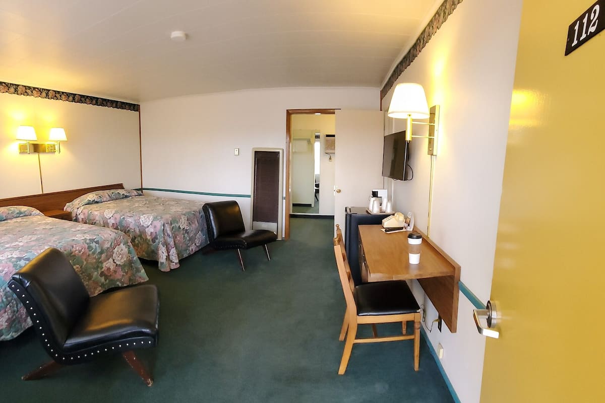 A hotel room with two beds, a desk, a chair, a TV, and a lamp.